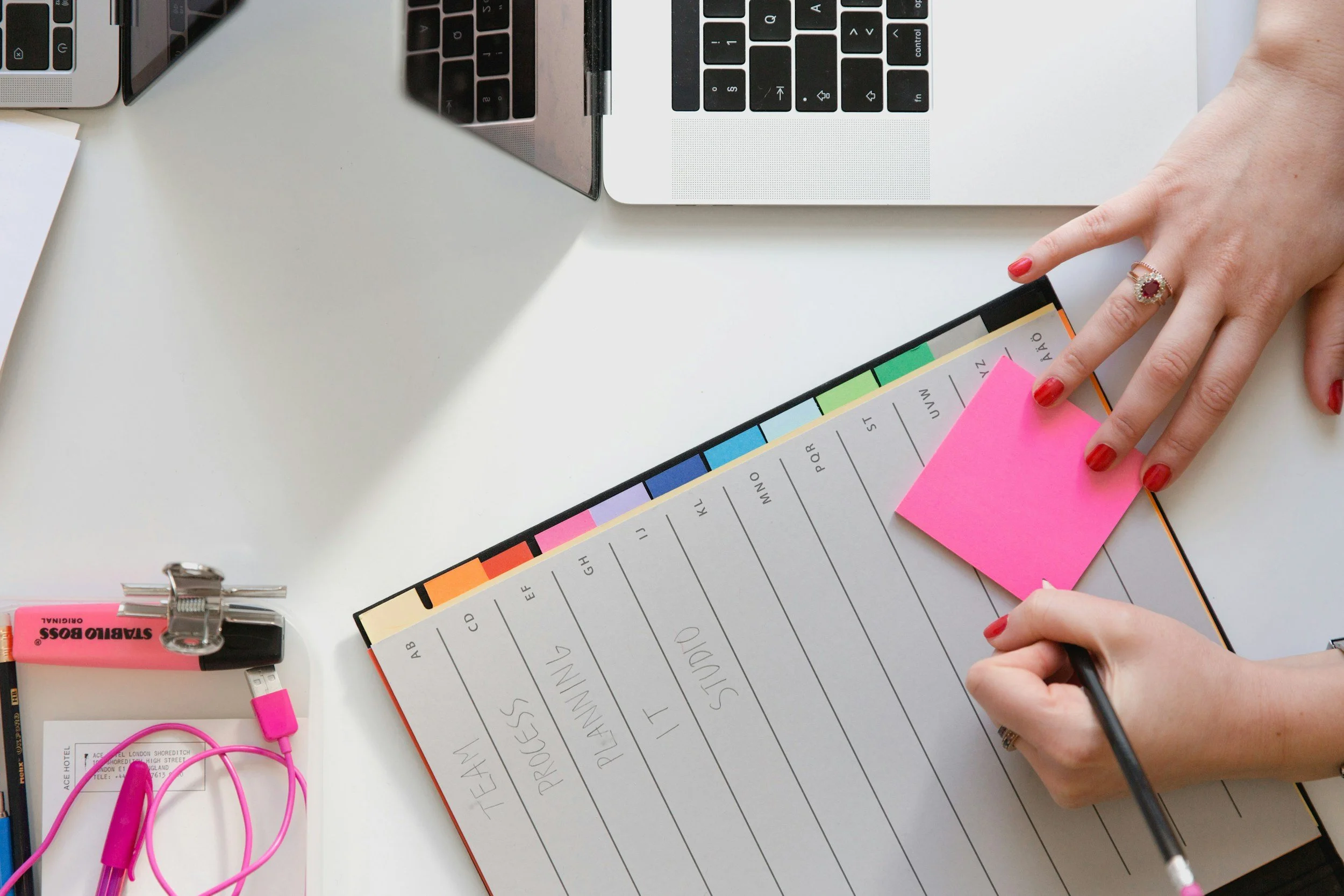 A person's hand writing on a planner with days of the week written in pencil. There is a pink sticky note on the planner and a person wearing a ring and red nail polish. Other office supplies include pink earbuds, a laptop, a pink highlighter, and a black clip.